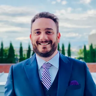 Bob Calise smiling outdoors in a navy suit and purple striped tie, with trees and blue sky behind him.